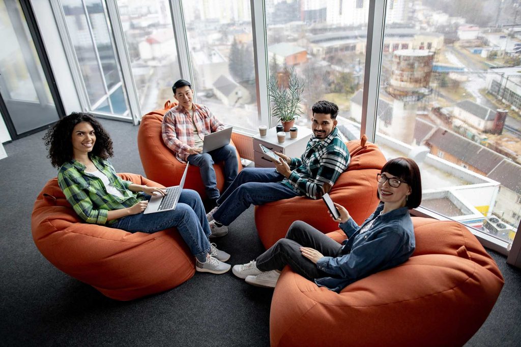 Top view of diverse male and female adults in casual clothes posing on terracotta soft seats with digital gadgets. Multiethnic collaborators planning presentation for colleagues in shared workspace.